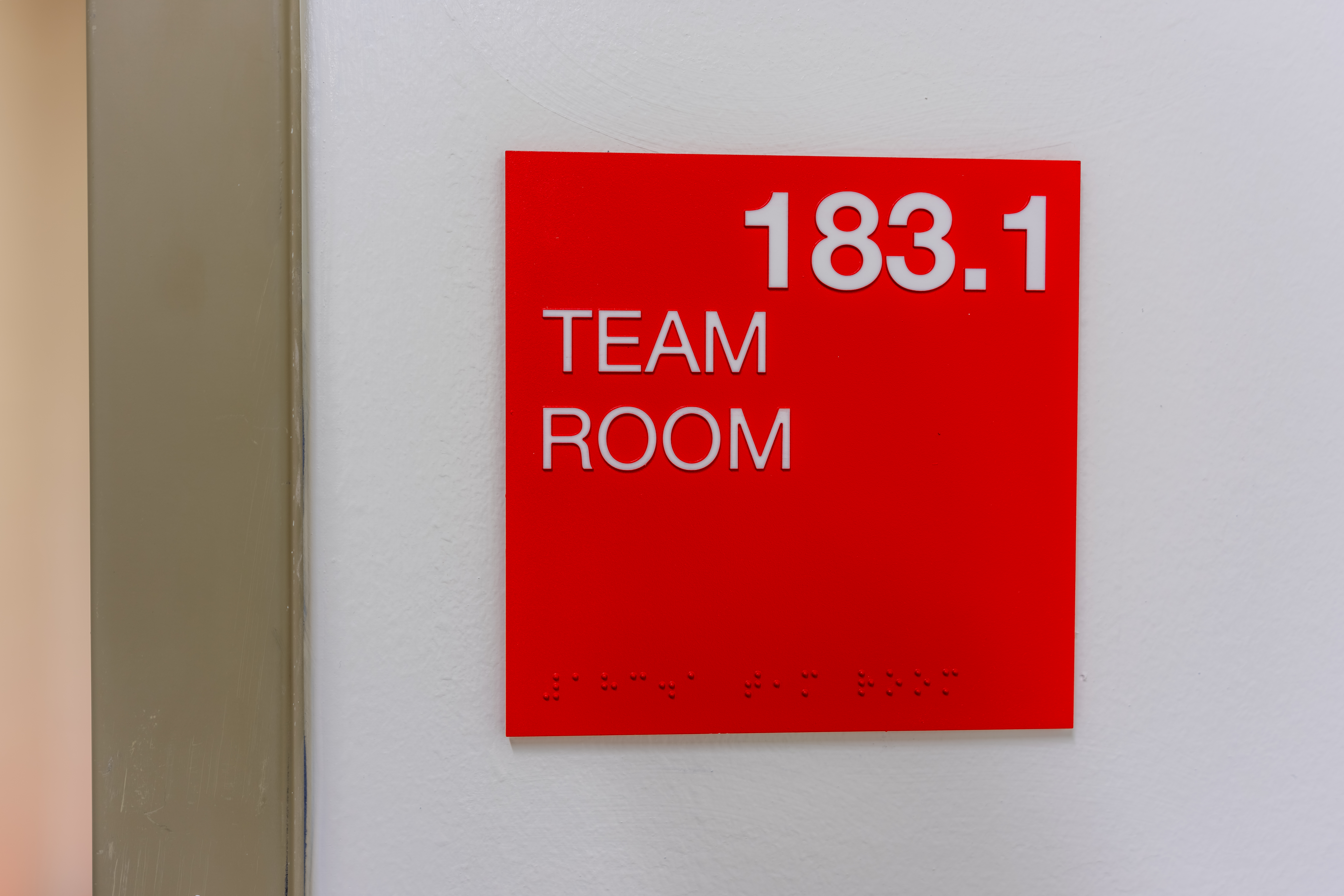 Red and white team room sign, 183.1, with braille code on a white interior building wall.